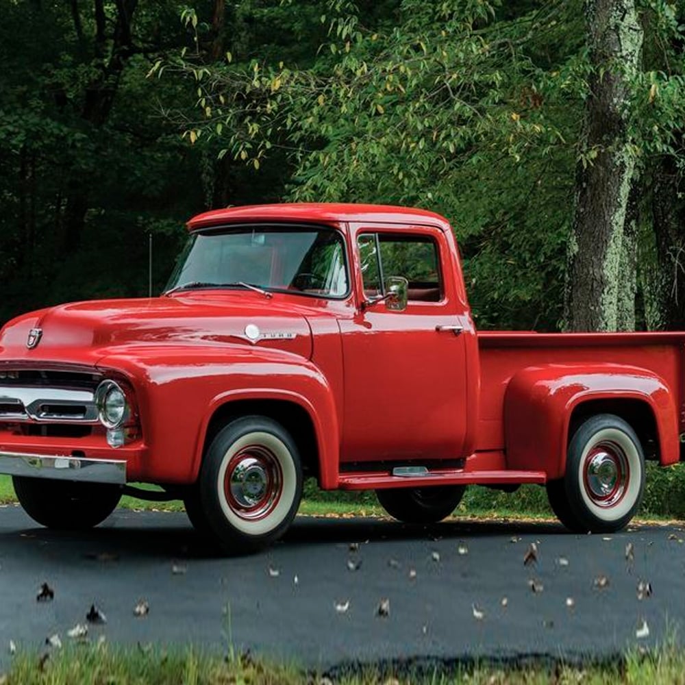 A bright red vintage pickup truck is parked on a paved driveway surrounded by green trees and grass, with fallen leaves scattered on the ground.