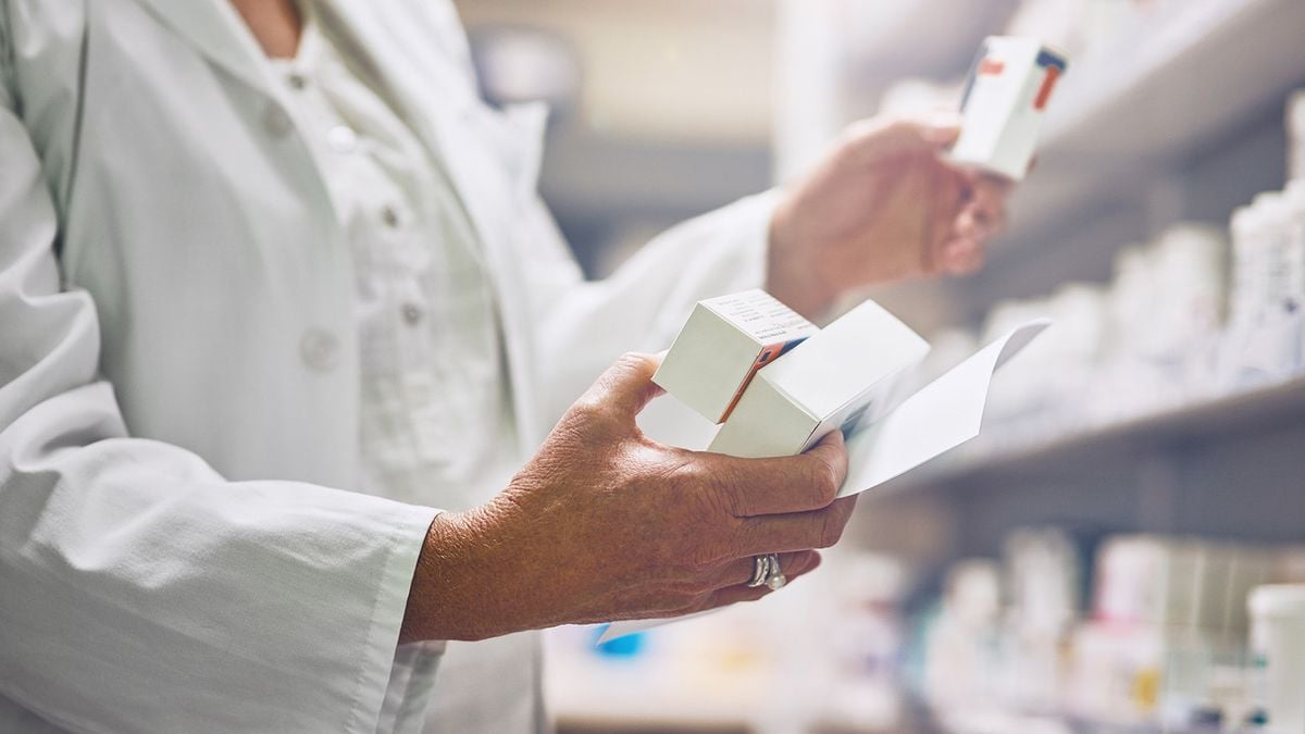 A pharmacist in a white coat holds and examines medication boxes in a pharmacy, with shelves of various medicines visible in the background.