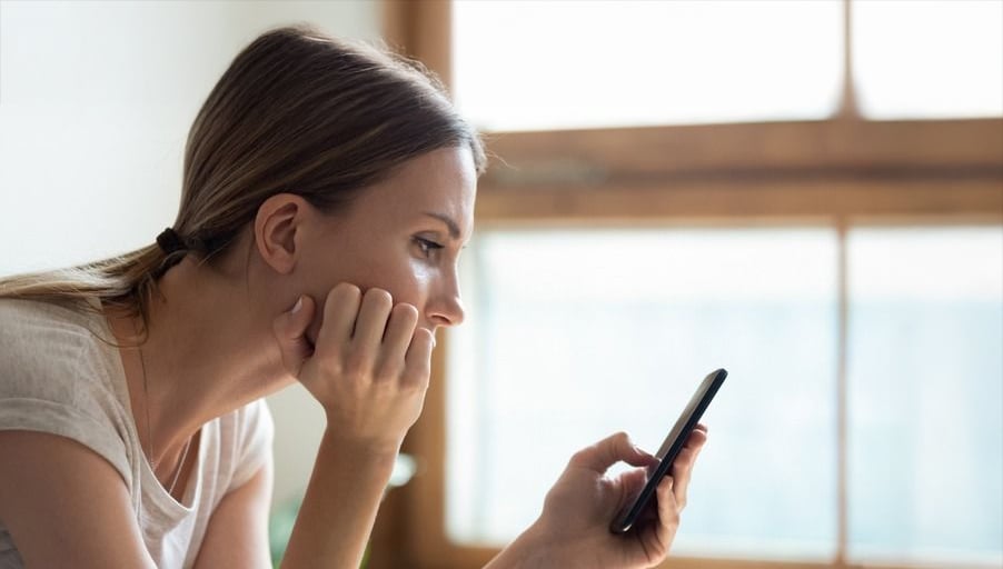 A woman sits indoors, resting her chin on her hand and looking intently at a smartphone she is holding, appearing thoughtful or concerned. A large window is in the background.