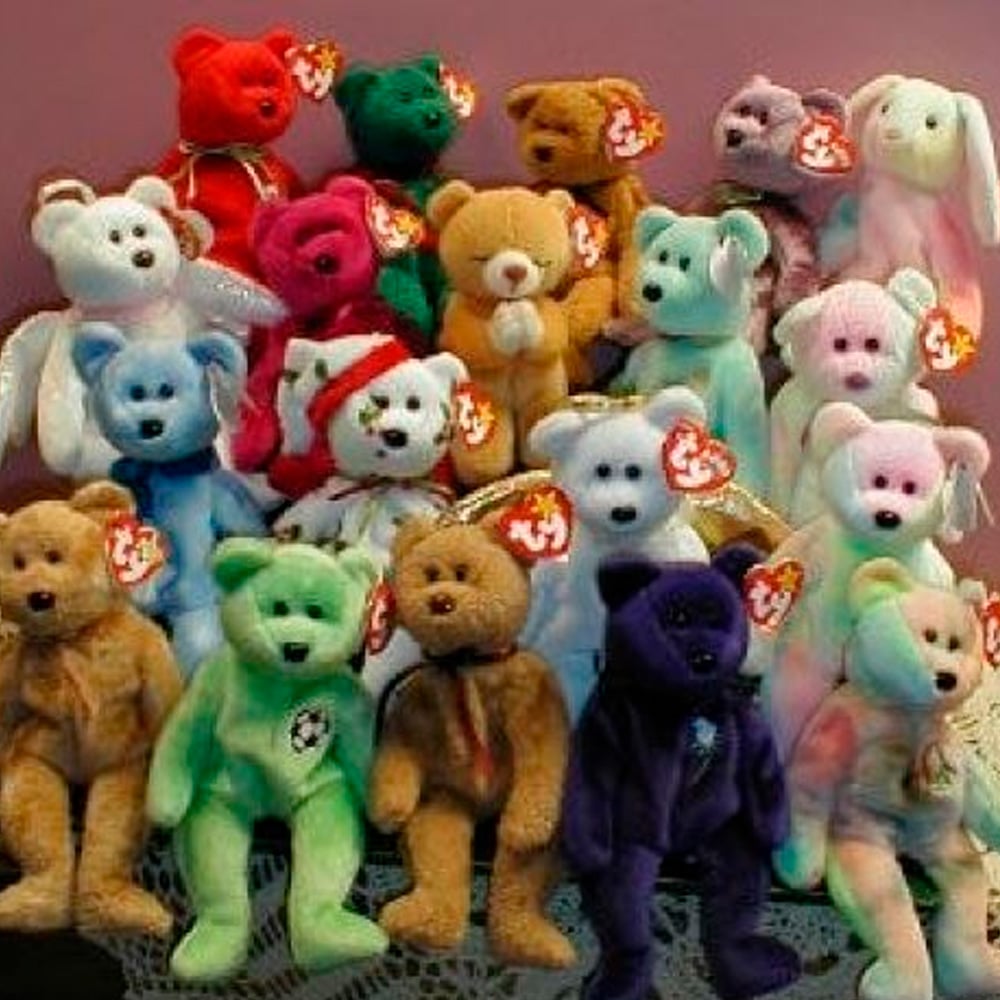 A collection of colorful Beanie Babies, including bears, a rabbit, and a dog, is arranged in three rows on a lace-covered surface. Each toy has a red and white Ty tag attached to its ear.