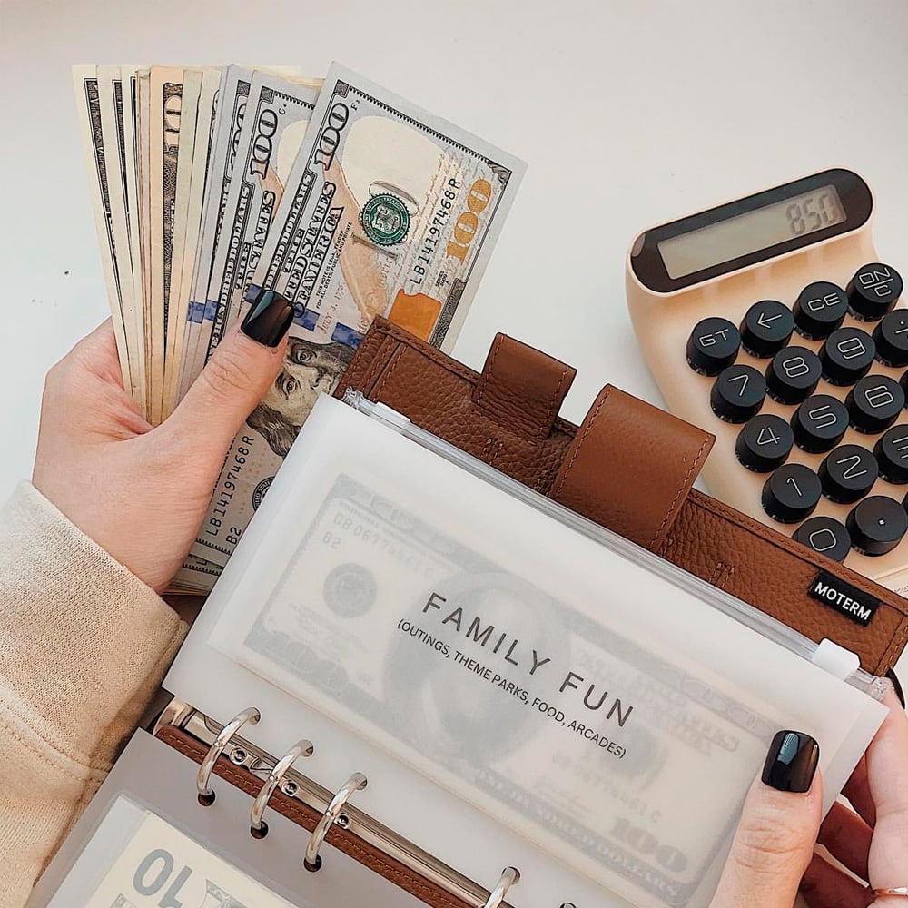 A person holding a stack of U.S. $100 bills and an open budget binder labeled "FAMILY FUN," with a beige calculator displaying 850 nearby.