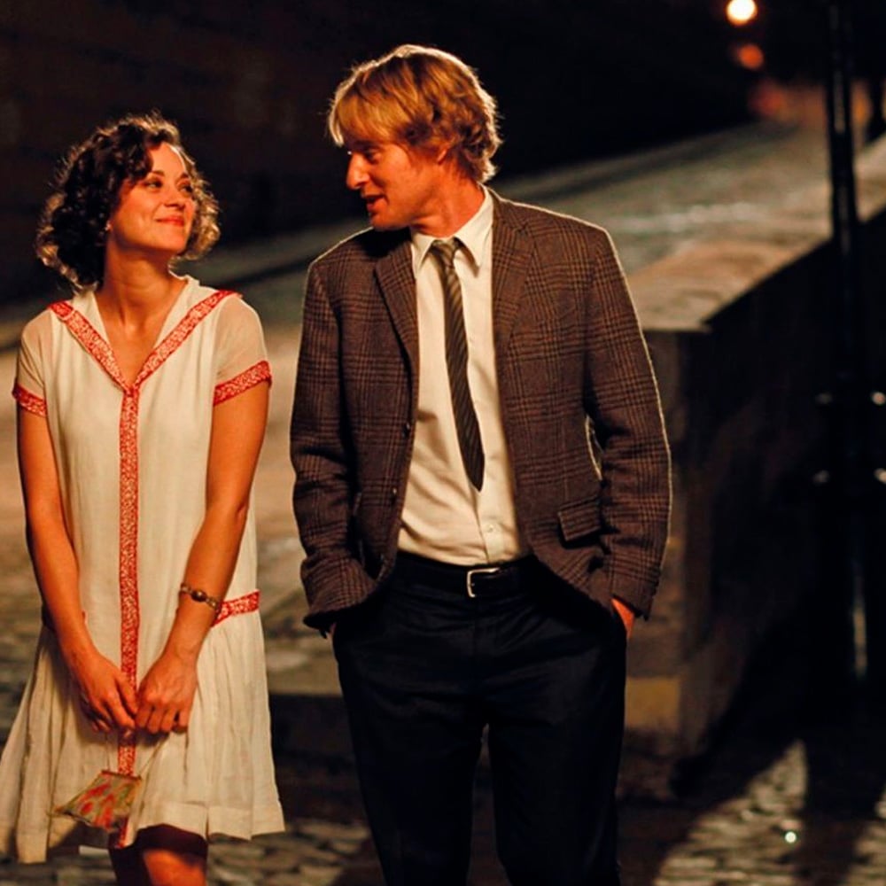 A woman in a white dress with red trim smiles at a man in a suit and tie as they walk together at night on a cobblestone street, illuminated by warm streetlights.