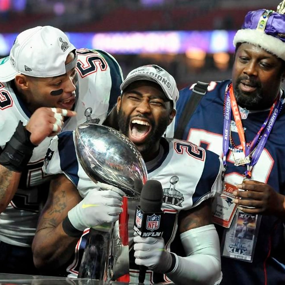 Three jubilant football players in New England Patriots uniforms celebrate with a Super Bowl trophy; one shouts joyfully while holding the trophy and a microphone, flanked by cheering teammates.