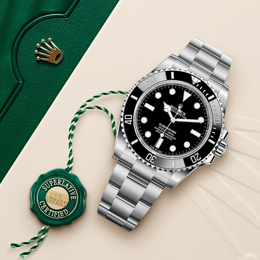A stainless steel Rolex Submariner watch with a black dial and bezel is displayed on a beige surface next to a green Rolex box, a gold Rolex crown logo, and a green "Superlative Certified" tag.