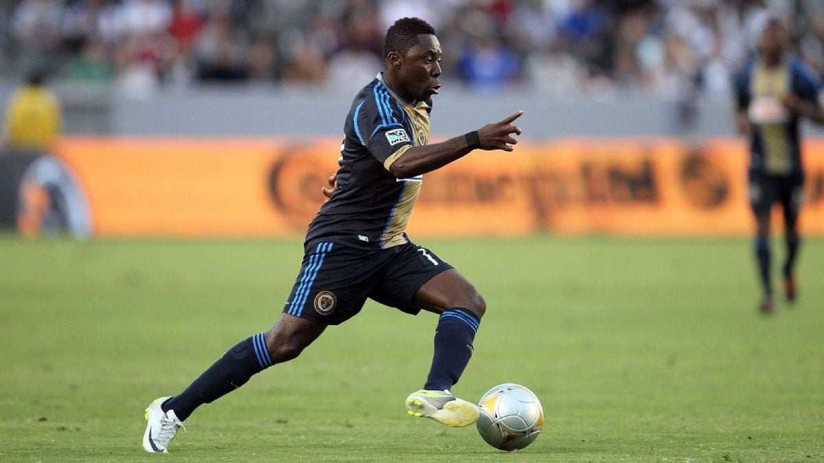 A soccer player wearing a dark blue and gold uniform runs with the ball on a grassy field during a match, with a blurred crowd and orange advertising boards in the background.
