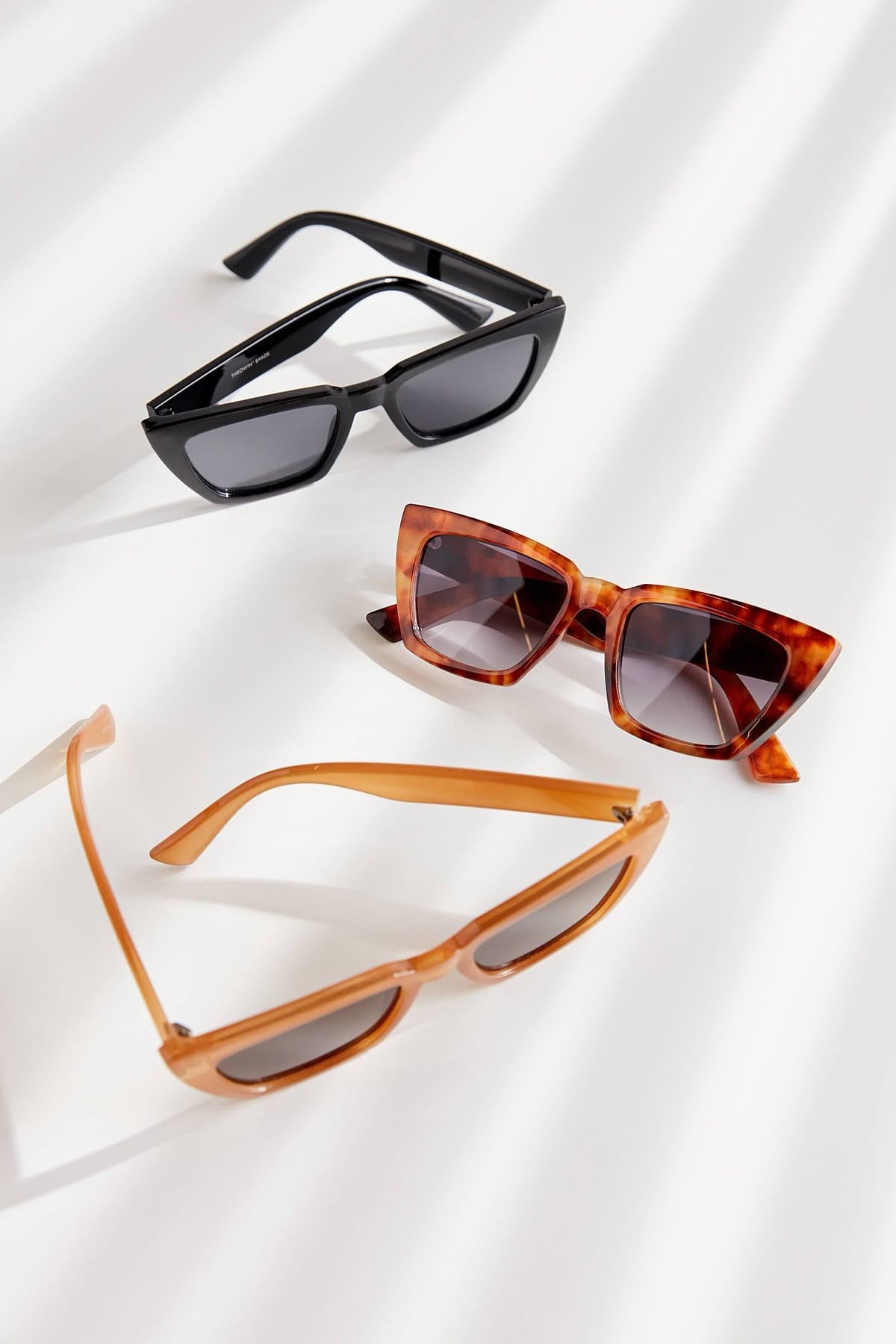 Three pairs of sunglasses are displayed on a white surface: one black with rectangular lenses, one brown with a tortoiseshell cat-eye frame, and one light tan with an oval frame. Shadows fall diagonally across the scene.