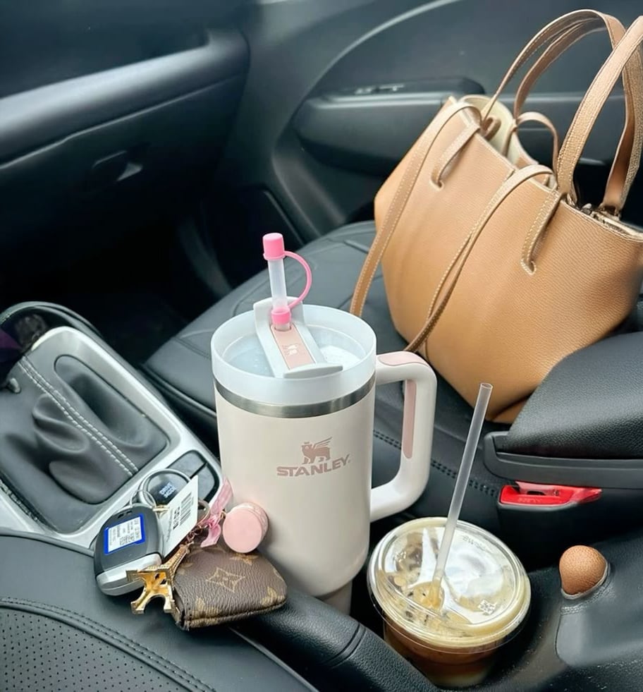 A car interior with a tan handbag on the passenger seat, a Stanley tumbler with a pink straw and lip balm, a set of car keys, a Blockbuster membership card, and an iced coffee in the cup holder.