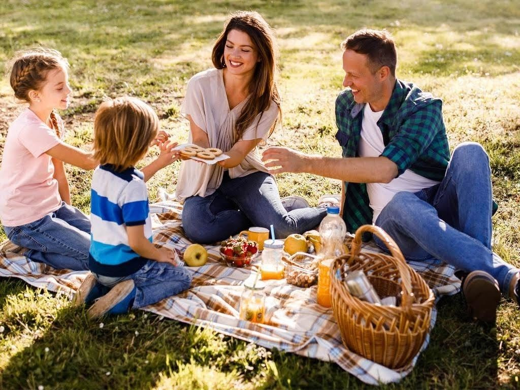 A family of four enjoys a picnic on a blanket in a sunny park, sharing food and smiling. A basket, fruits, and drinks are visible around them as they sit on the grass.