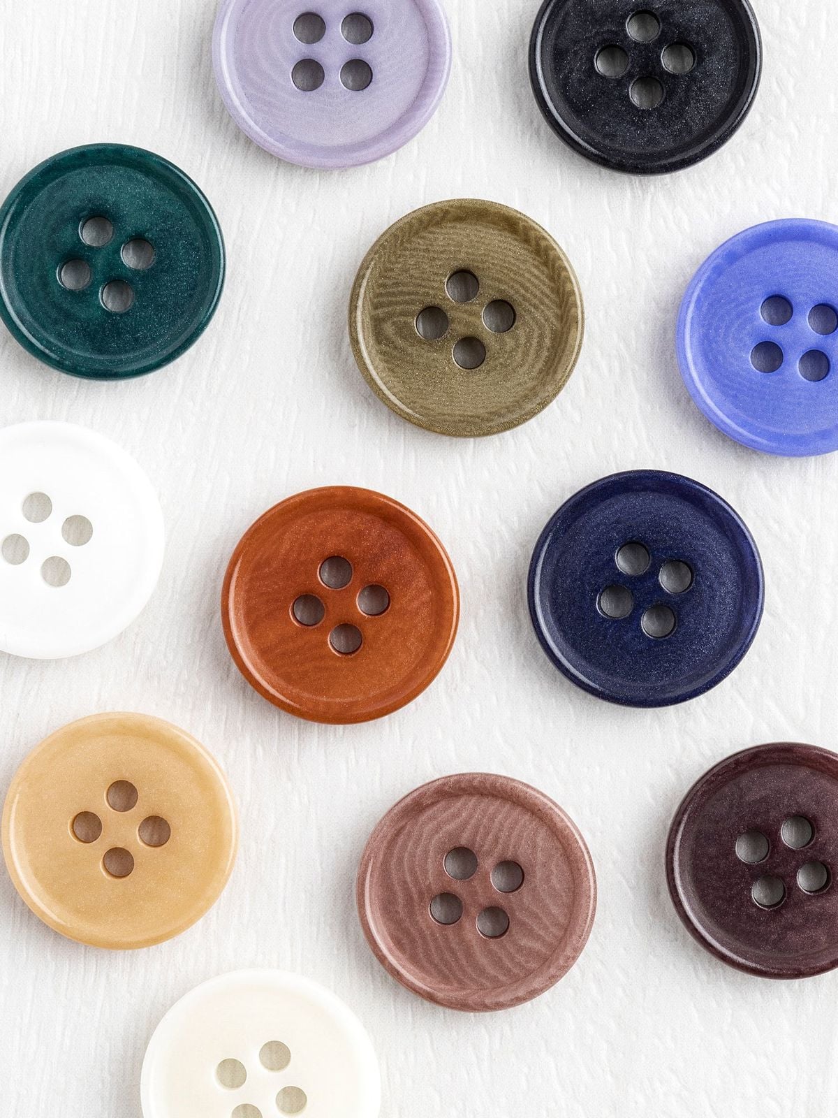 A variety of round, four-holed buttons in different colors—green, purple, black, white, brown, navy, beige, and blue—arranged in a scattered pattern on a white textured surface.