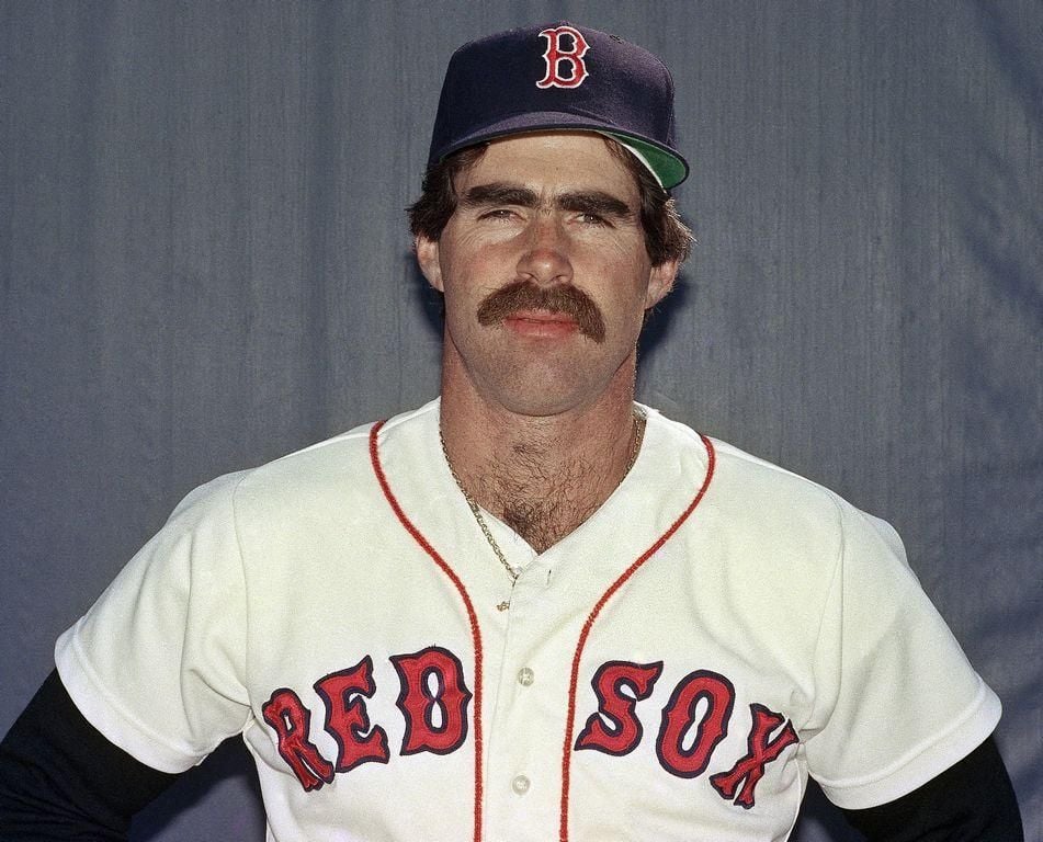 A baseball player in a Boston Red Sox uniform and cap stands against a plain background, looking at the camera with a neutral expression and sporting a thick mustache.