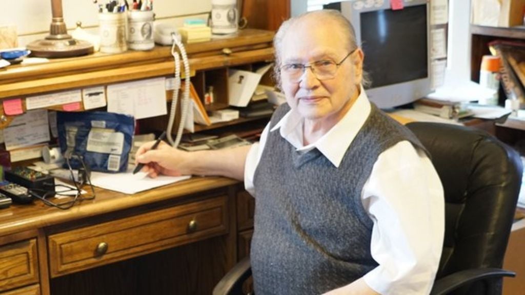 An older man with glasses and a gray sweater vest sits at a wooden desk covered with papers and office supplies, looking at the camera and holding a pen. Shelves and a computer monitor are in the background.