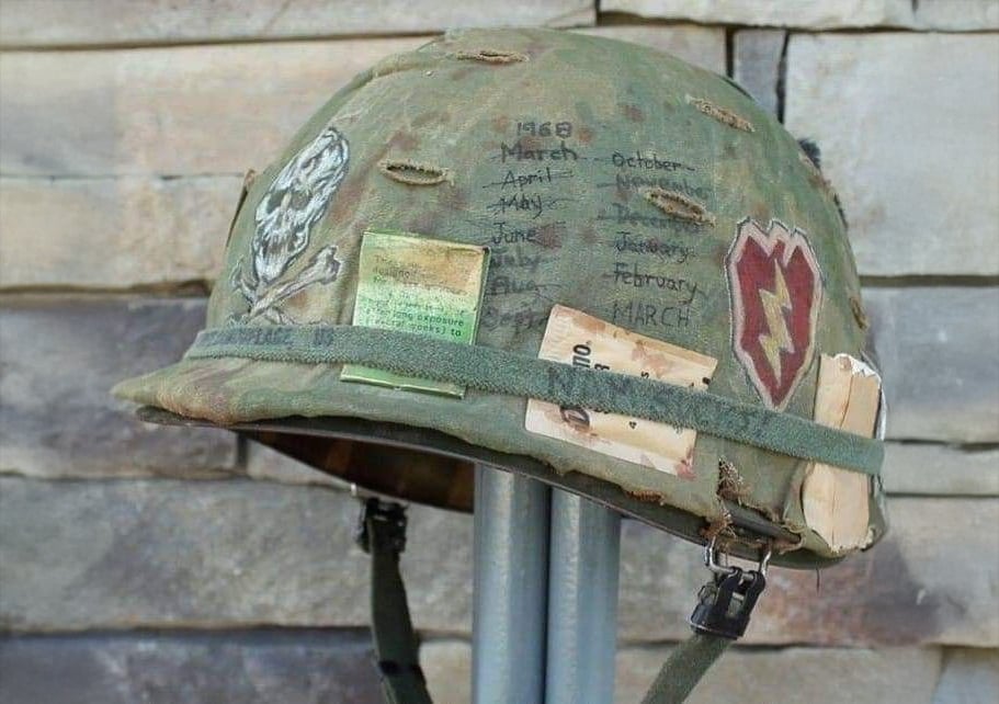 A worn military helmet with handwritten dates, faded patches, a skull drawing, and visible signs of damage, hanging on a stand in front of a stone wall background.