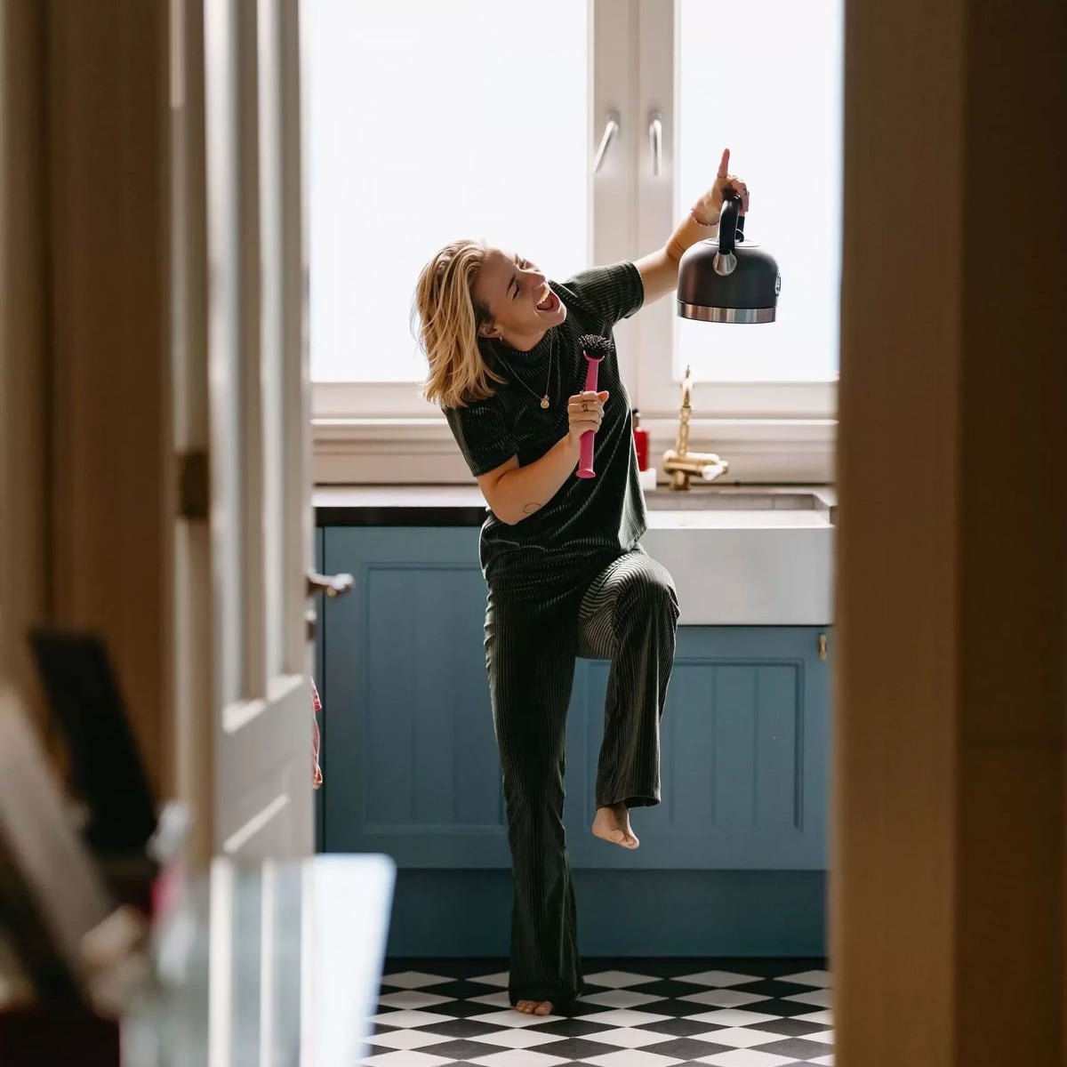 A woman in pajamas dances joyfully in a kitchen, holding a kettle in one hand and a pink object in the other, with one foot raised and a big smile on her face.