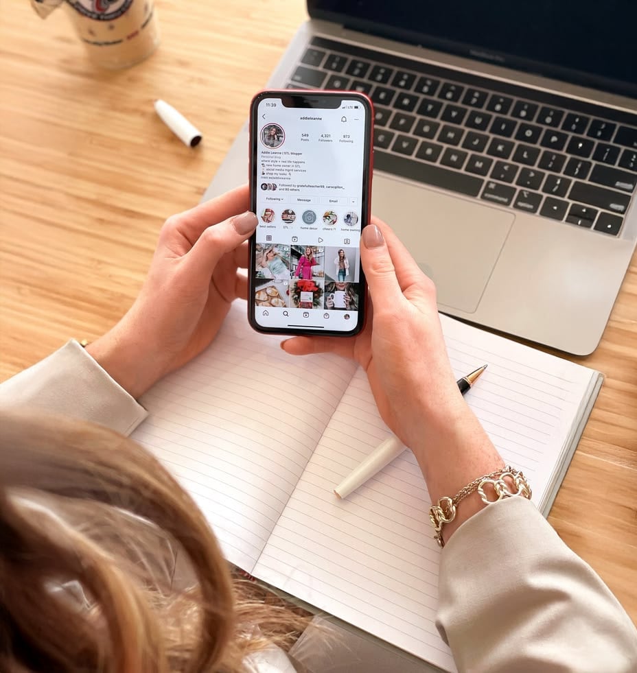 A person sits at a desk with a laptop, open notebook, and pen, holding a smartphone displaying an Instagram profile page. A coffee cup is also on the desk.