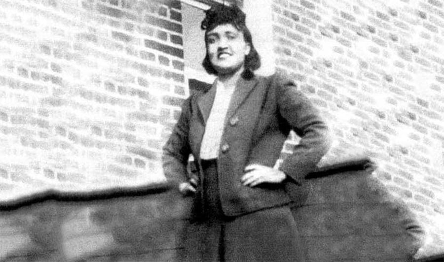 A Black woman in a dark suit stands confidently outside in front of a brick wall, with her hands on her hips, looking slightly up and smiling.