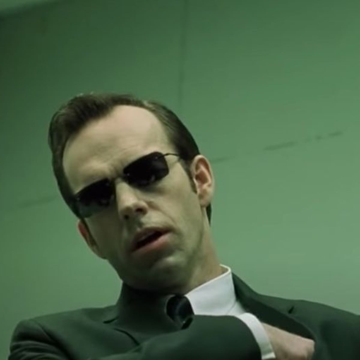 A man with slicked-back hair wearing dark sunglasses, a suit, and a tie stands against a green-tinted background, looking stern and serious.