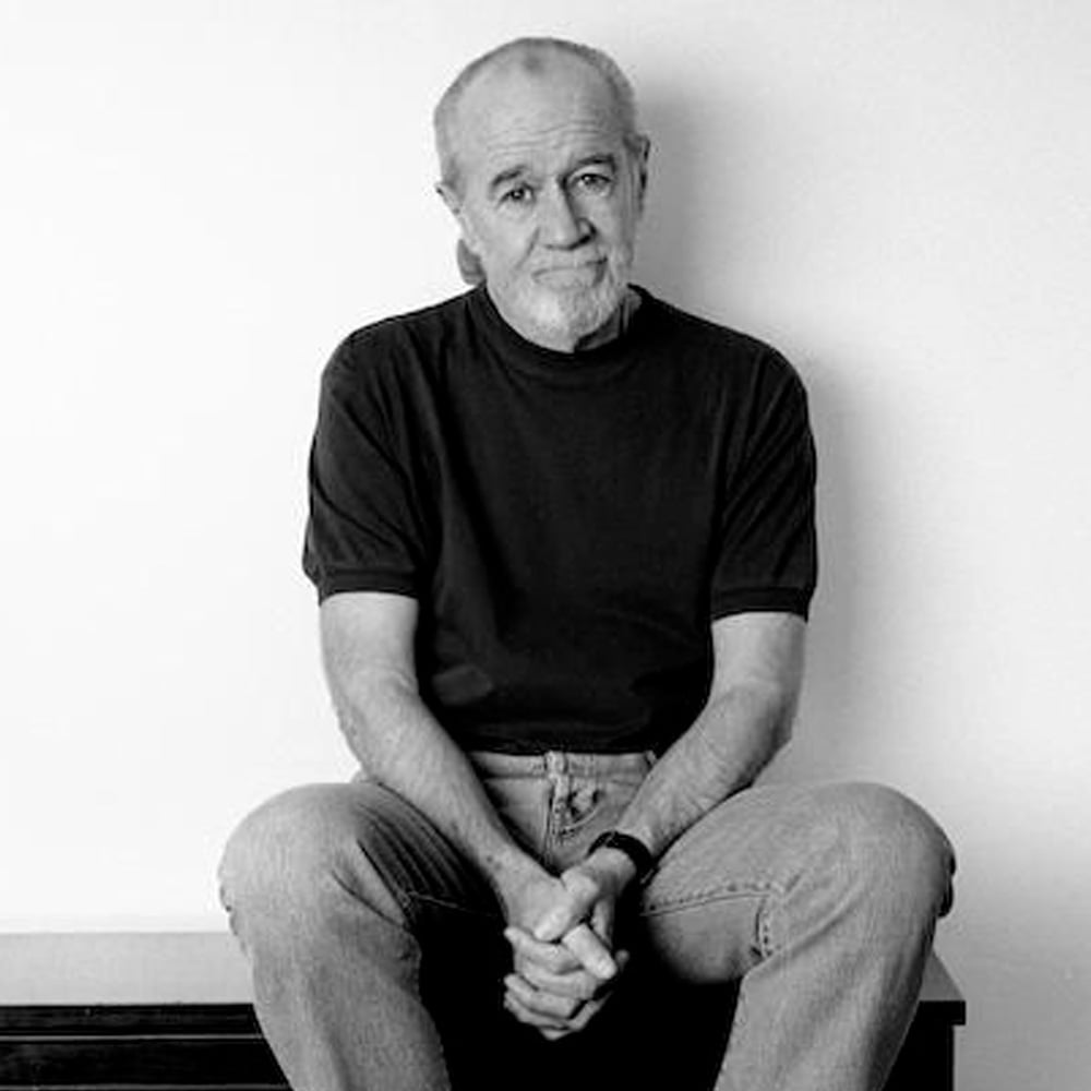 A man with short gray hair and a beard sits on a bench against a plain white wall, wearing a dark t-shirt and light jeans, with his hands clasped loosely in front of him and a thoughtful expression on his face.