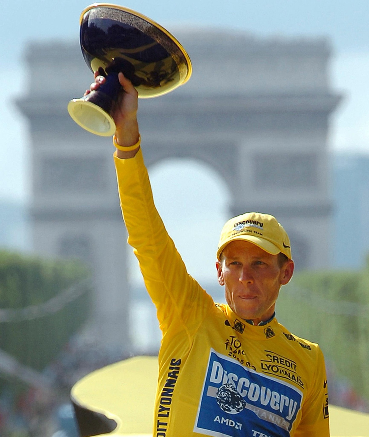 A cyclist in a yellow jersey and cap holds a trophy aloft in front of the Arc de Triomphe in Paris, celebrating a victory. The crowd and avenue are visible in the background.