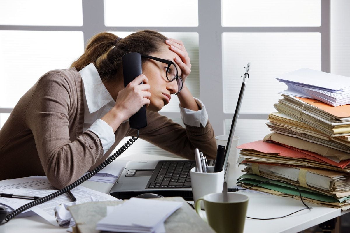A stressed woman wearing glasses holds a phone to her ear and rests her head on her hand while sitting at a cluttered desk with a laptop, paperwork, folders, and coffee cups.