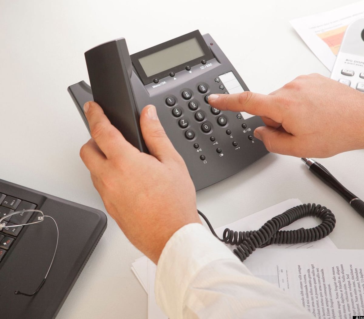 A person in a white shirt uses one hand to hold a telephone receiver and the other hand to dial a number on a desk phone, with papers and a laptop nearby on the desk.