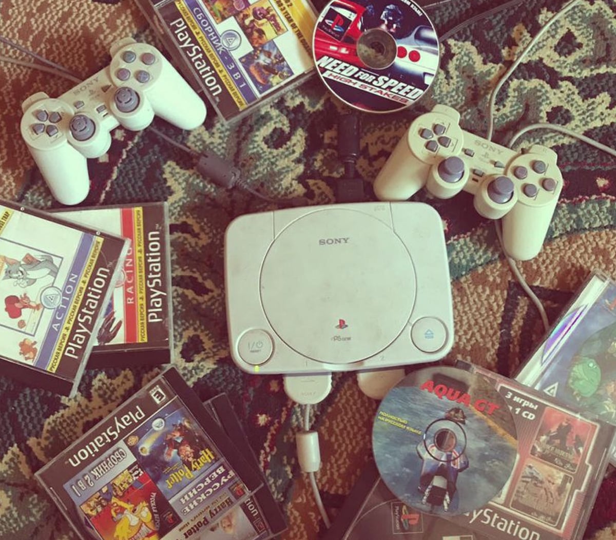 A white Sony PlayStation console with two controllers is surrounded by various PlayStation game cases and discs on a patterned rug. Games include Need for Speed, Aquanaut's Holiday, and others.