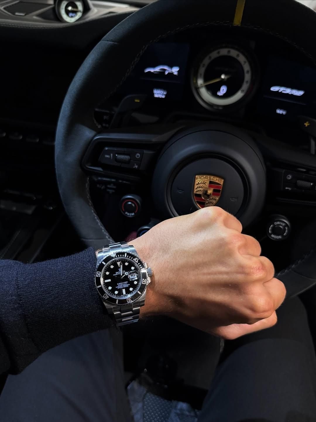 A person’s wrist wearing a silver luxury watch is held up in front of a steering wheel with the Porsche logo and a digital dashboard inside a sports car.