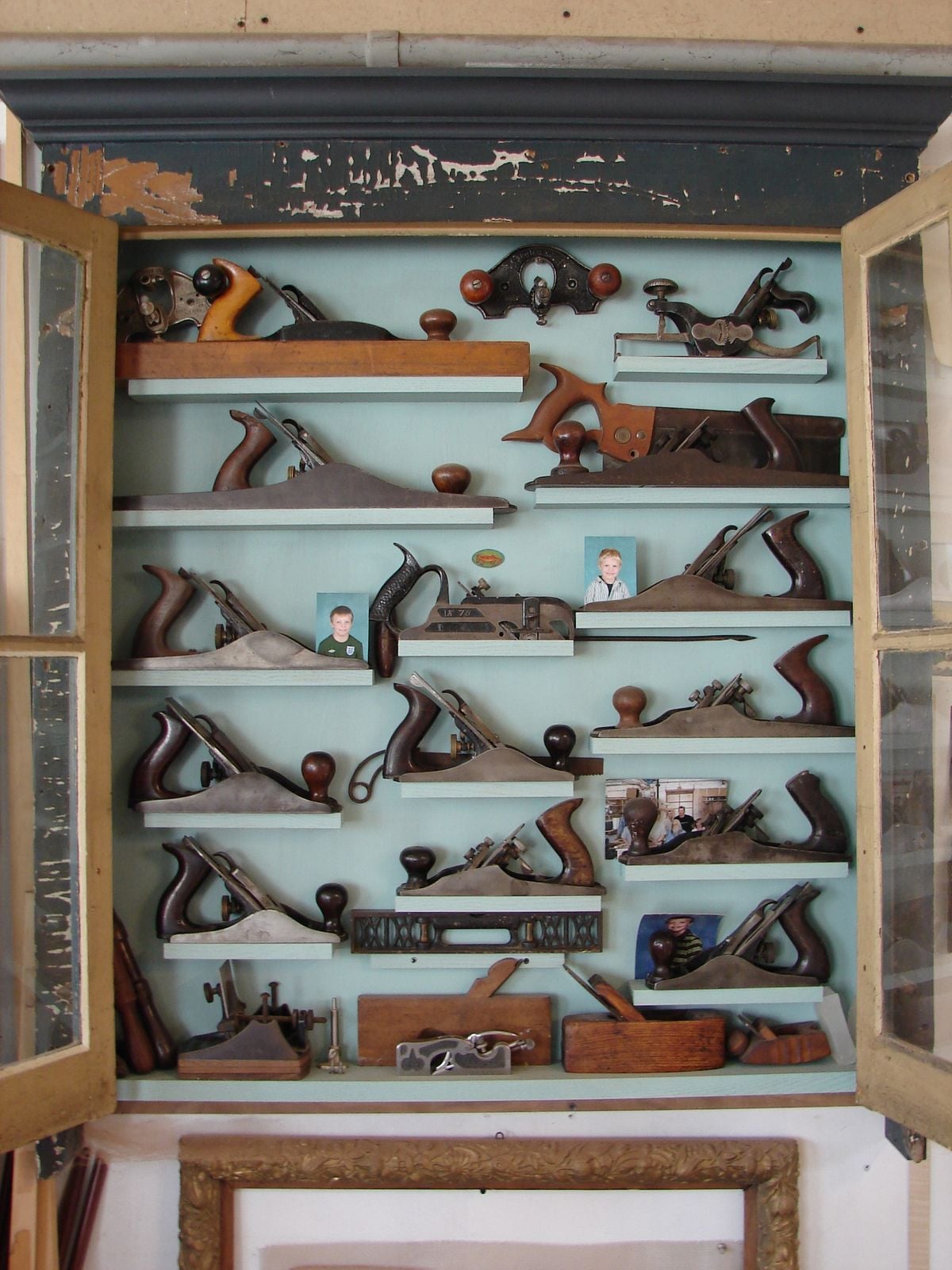 A wooden cabinet with open glass doors displays various vintage hand planes arranged on shelves, along with a few small photographs and woodworking tools.