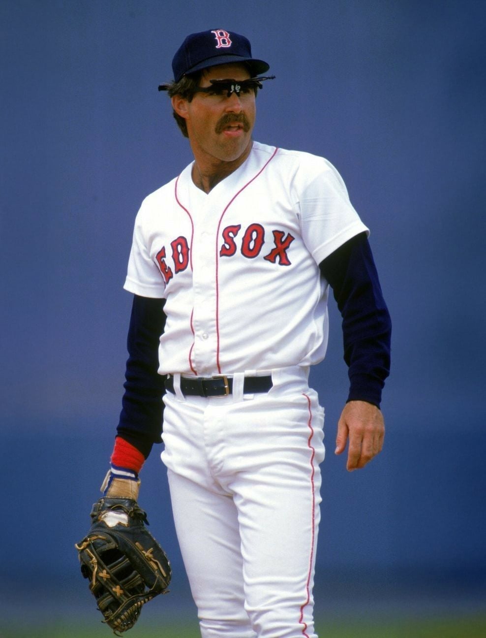 A baseball player in a Boston Red Sox uniform stands on the field with a glove on his left hand, wearing sunglasses and a cap, against a plain blue background.