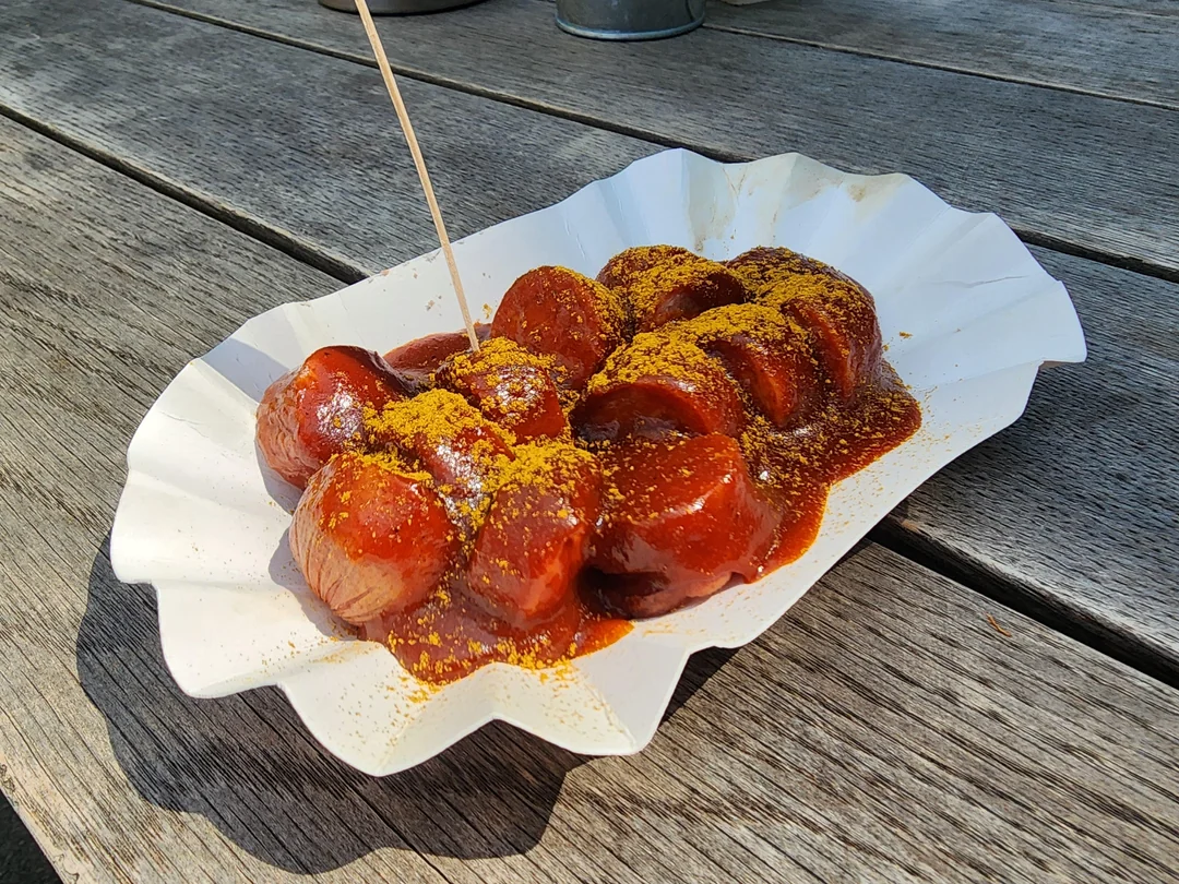 A paper tray filled with sliced sausage covered in reddish sauce and yellow curry powder, with a wooden pick, on a wooden table in sunlight.