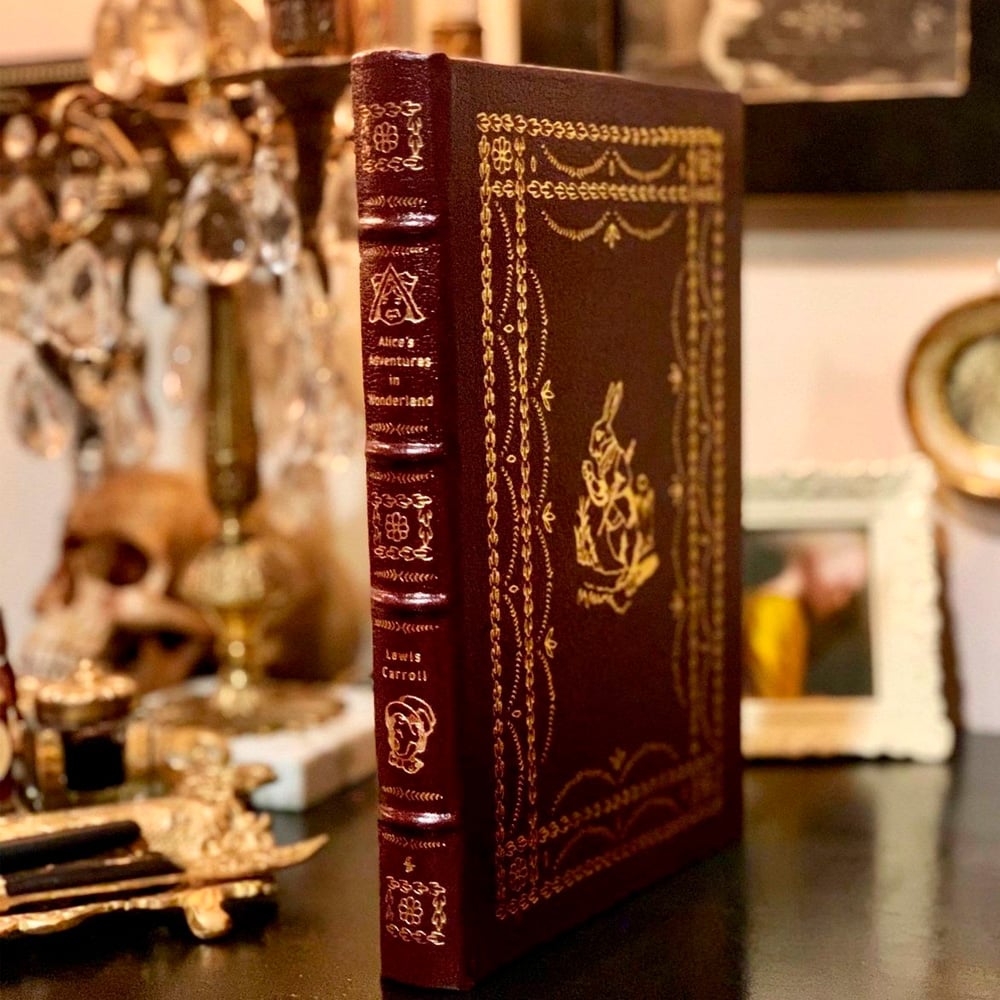 A richly decorated, leather-bound edition of "Alice's Adventures in Wonderland" stands upright on a cluttered surface with vintage décor, including a framed photo, a candlestick, and a skull in the background.