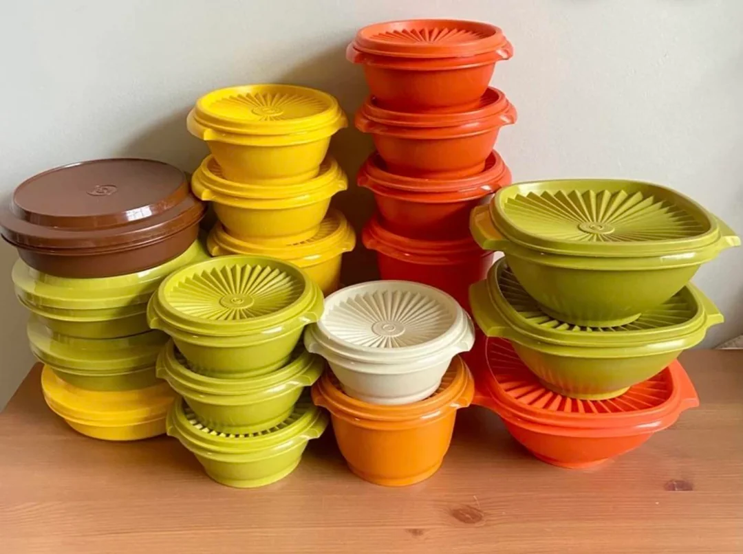 A collection of colorful vintage Tupperware containers in orange, yellow, green, beige, and brown, stacked neatly on a wooden surface against a plain wall.