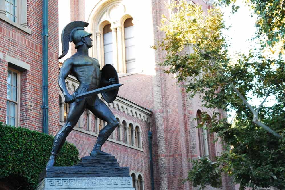 A bronze statue of a Trojan warrior with a helmet, shield, and sword stands in front of a red brick building with arched windows and leafy trees.