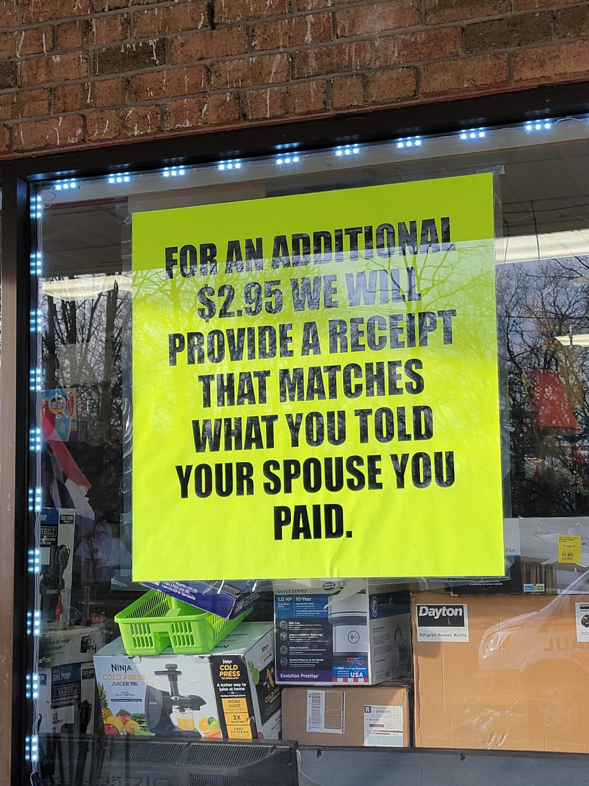 A bright yellow sign on a store window reads: “For an additional $2.95 we will provide a receipt that matches what you told your spouse you paid.” Store merchandise is visible through the window.