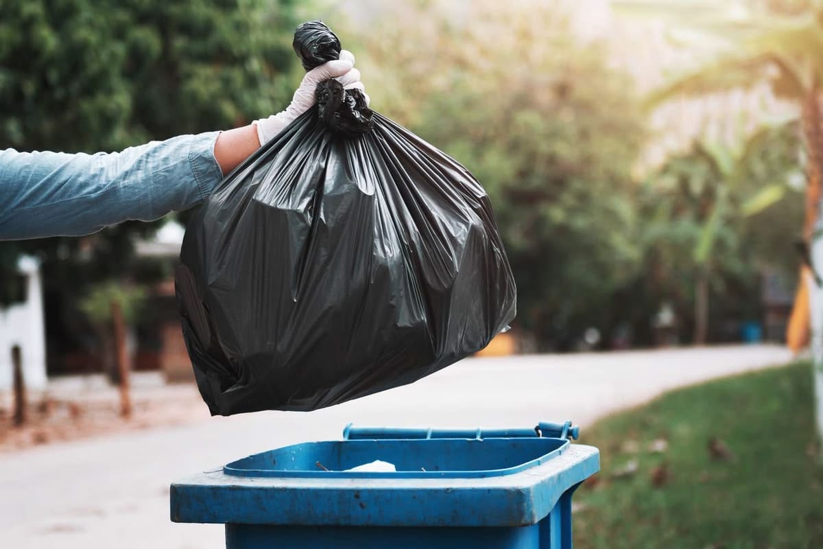 A person wearing a glove holds a black garbage bag over a blue trash bin outdoors, preparing to dispose of the bag. Trees and a road are visible in the blurred background.