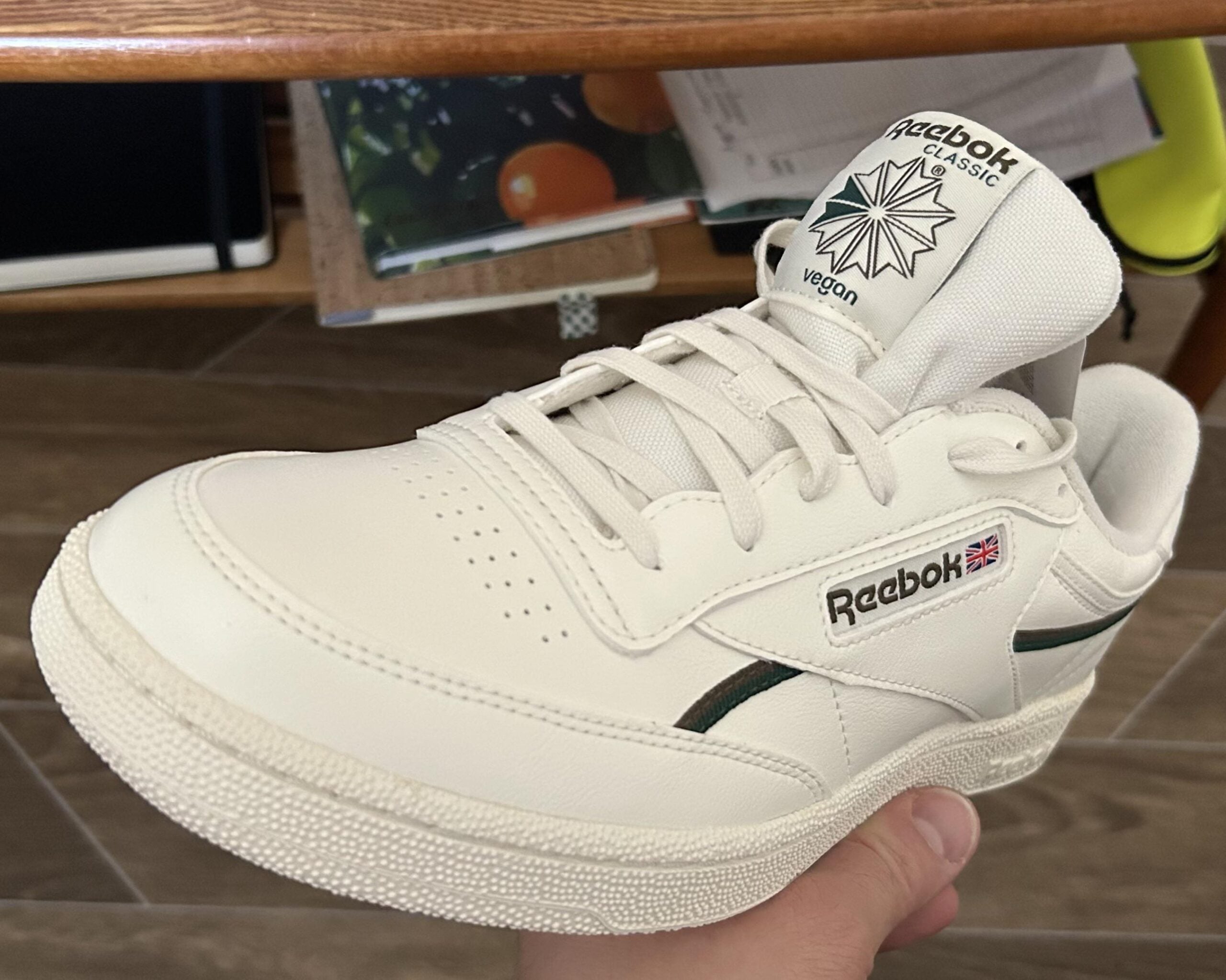 A hand holds a white Reebok Classic vegan sneaker with green accents, featuring the Reebok logo and a Union Jack flag on the side and “vegan” text on the tongue. Brown wooden shelves with books are in the background.