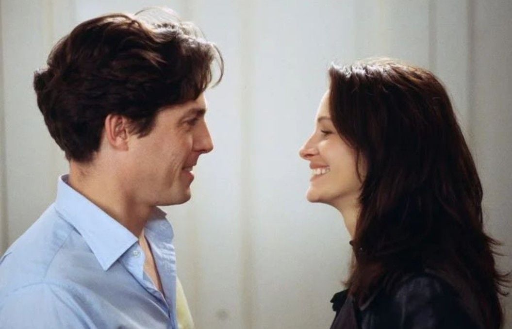 A man and a woman stand facing each other, smiling warmly. The man has short brown hair and wears a light blue shirt, while the woman has long brown hair and wears a dark jacket. They appear happy and relaxed in a softly lit room.