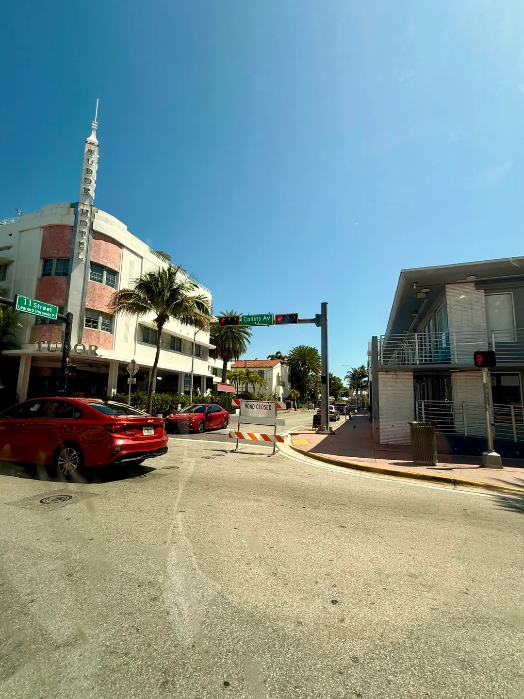 A sunny street scene in Miami Beach shows colorful art deco buildings, palm trees, a red car, and a "Road Closed" barricade at an intersection with a bright blue sky overhead.
