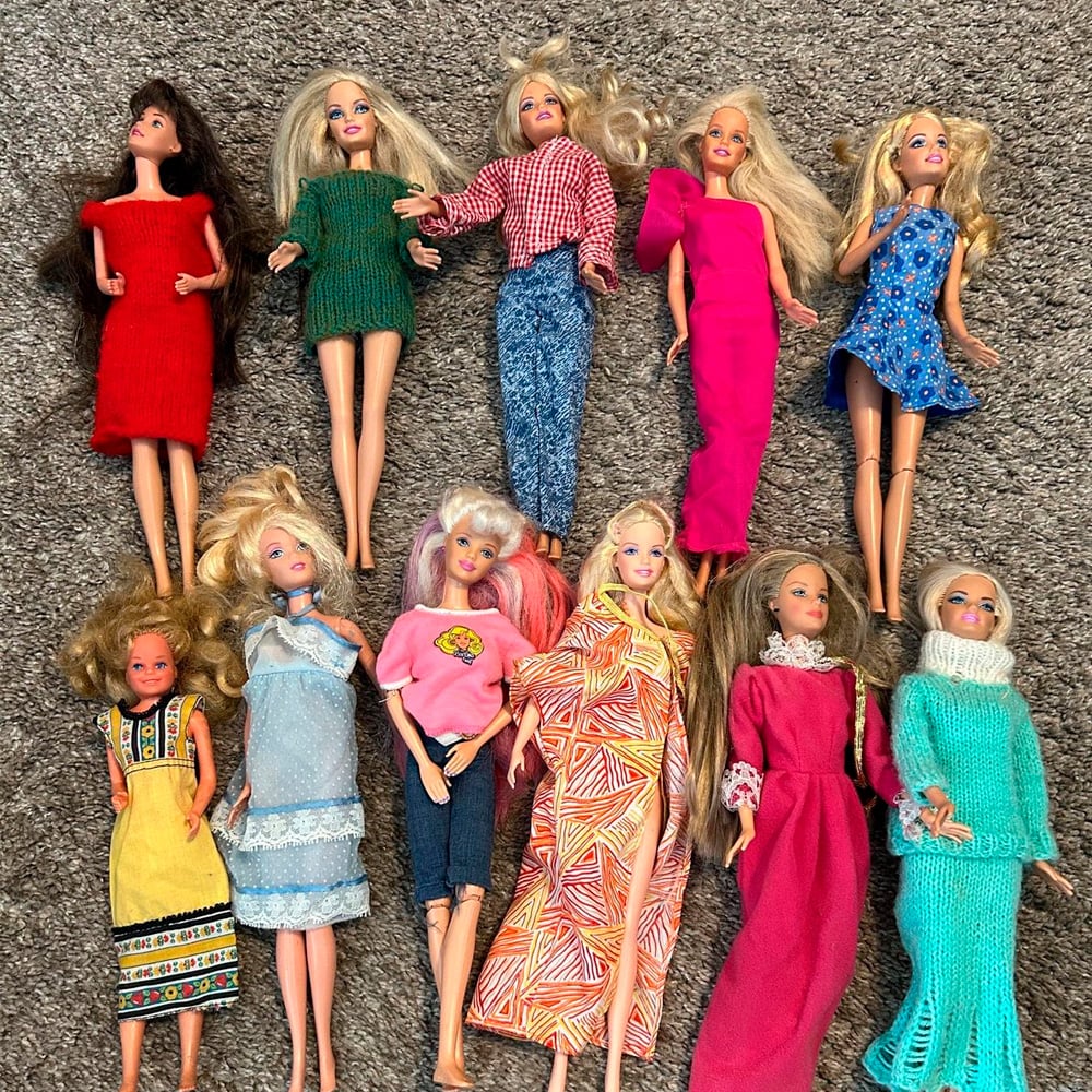 Ten Barbie dolls with various long hairstyles and colorful outfits are arranged on a gray carpet. Their clothes include dresses, jeans, skirts, and sweaters in bright and patterned fabrics.