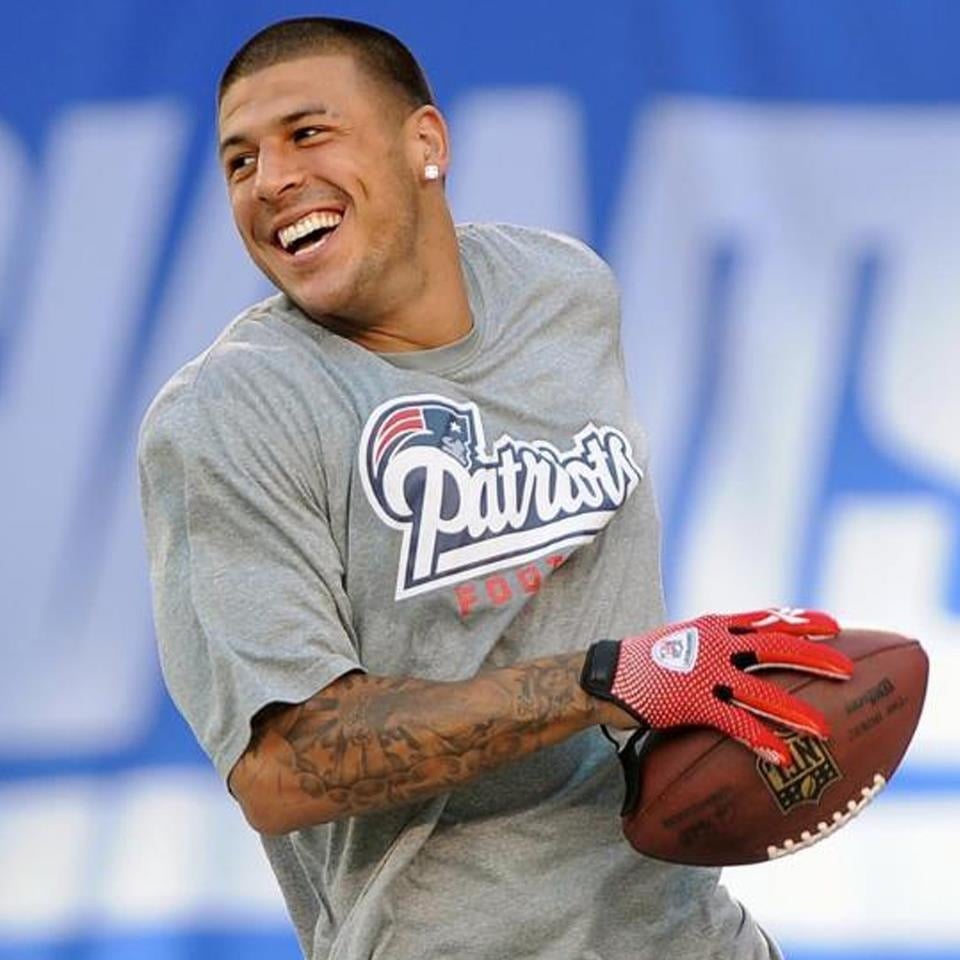 A smiling football player wearing a grey New England Patriots shirt and red gloves holds a football while looking over his shoulder on a field.
