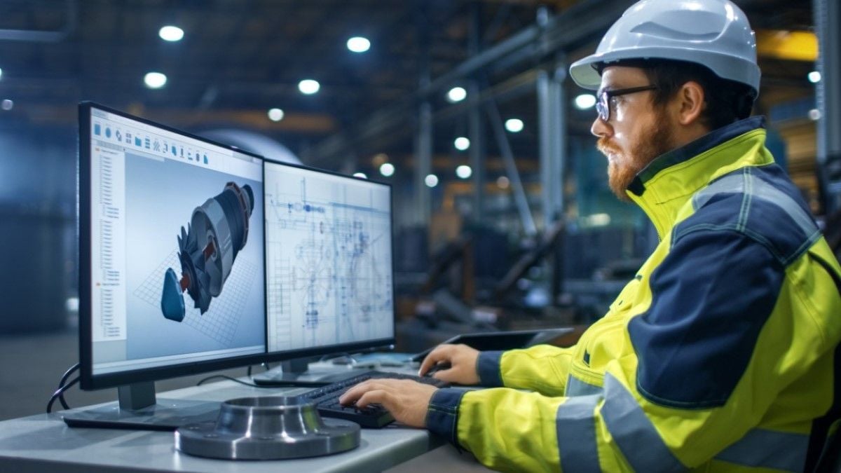 A male engineer wearing a hard hat and high-visibility jacket works at a desk with dual monitors displaying 3D mechanical designs and technical schematics in an industrial setting.