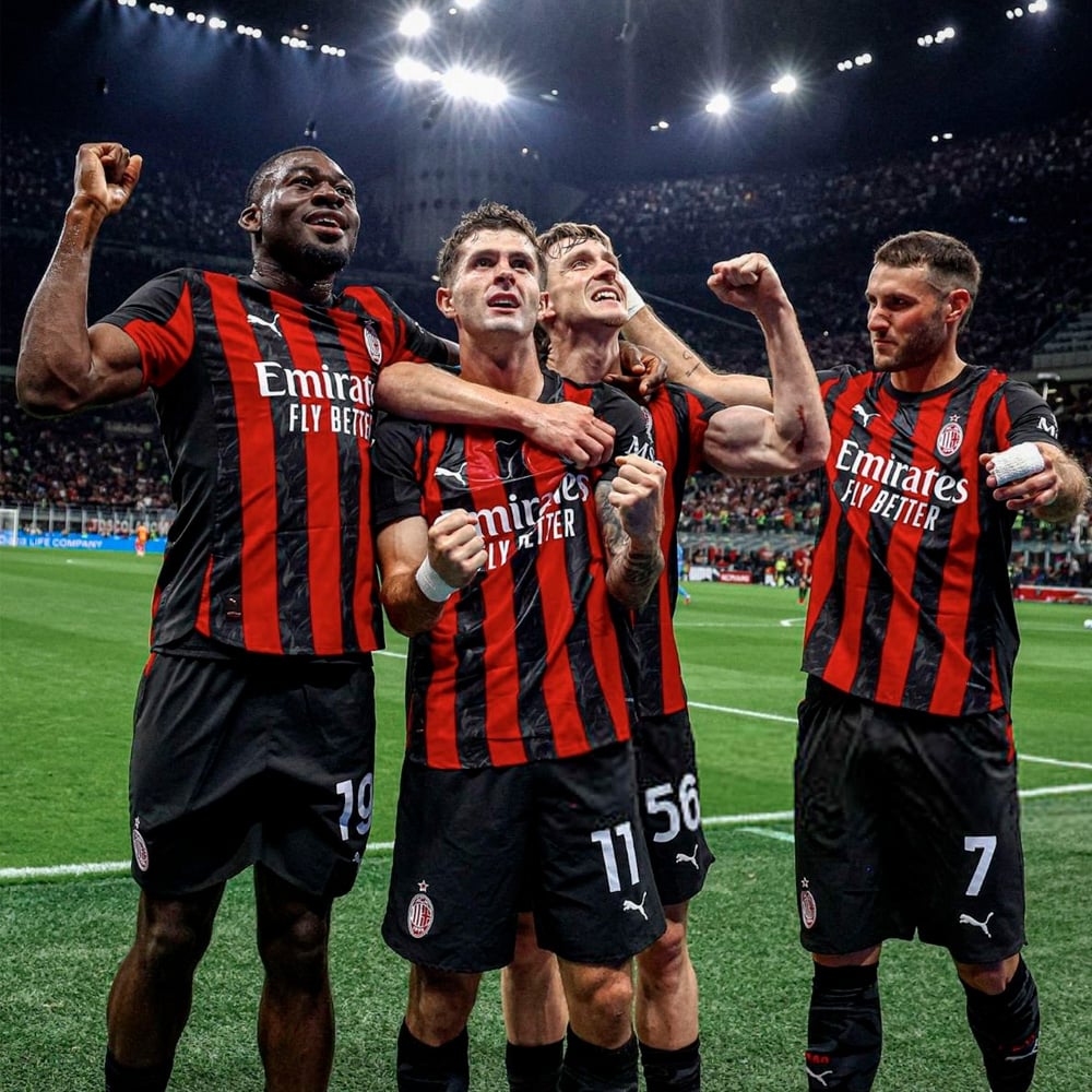 Four AC Milan soccer players wearing red and black striped uniforms celebrate on the field, raising their fists in triumph with a stadium full of fans in the background.