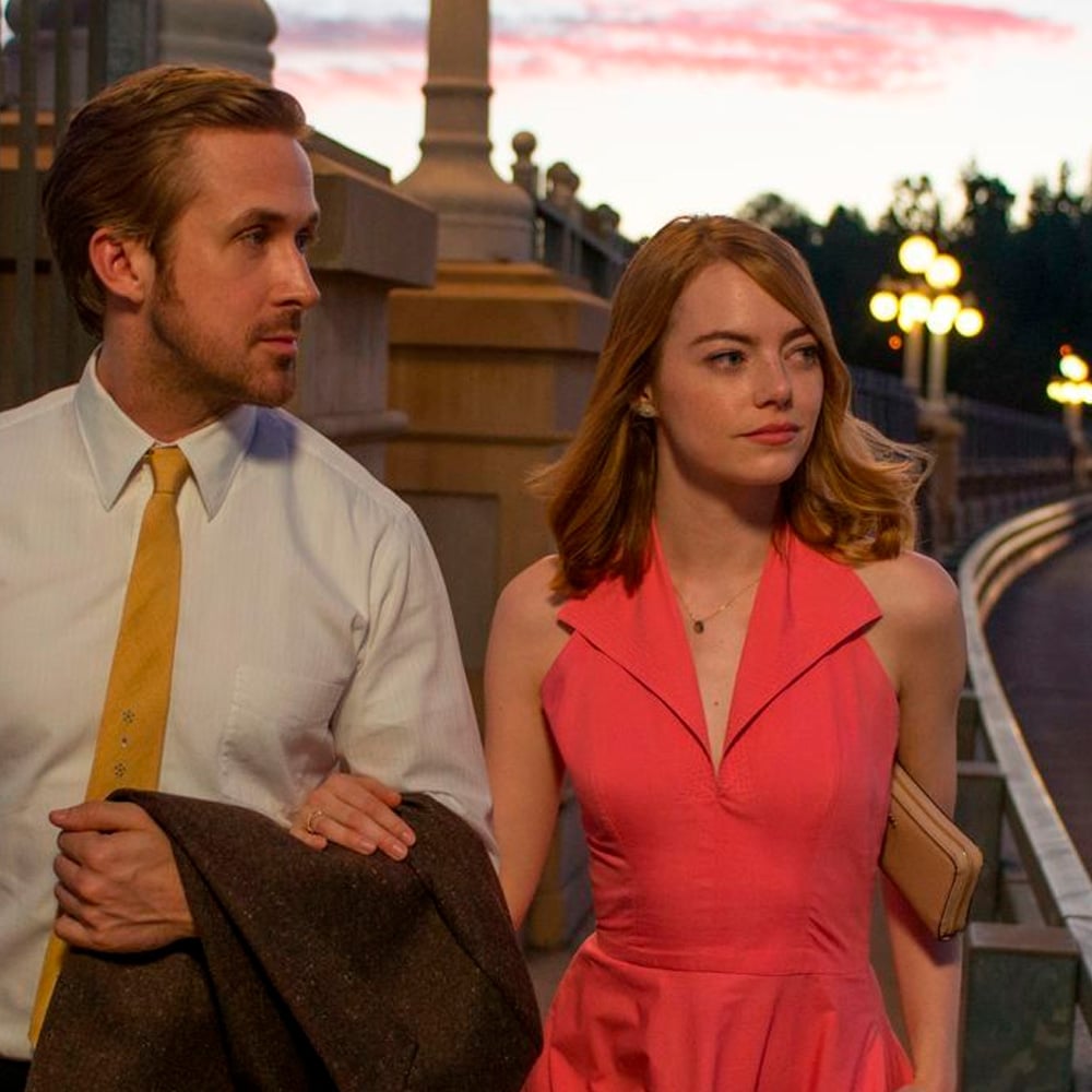 A man in a white shirt and yellow tie walks beside a woman in a sleeveless pink dress at sunset, holding her arm as they stroll along a bridge with lit lamps.