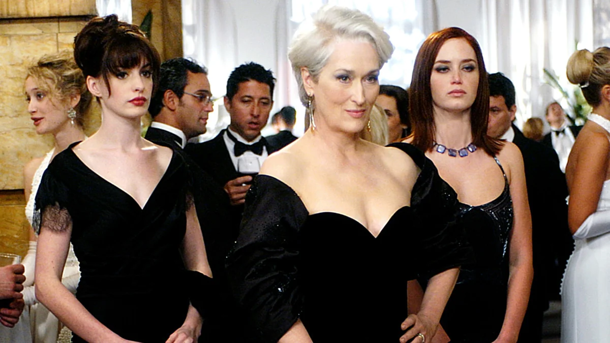 Three elegantly dressed women in black evening gowns stand together at a formal event, surrounded by people in suits and gowns in a sophisticated, well-lit room.