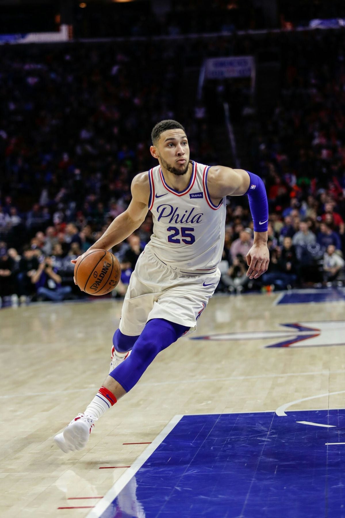 A basketball player in a white Philadelphia 76ers uniform dribbles the ball on a court, appearing to drive toward the basket during a game, with spectators visible in the background.