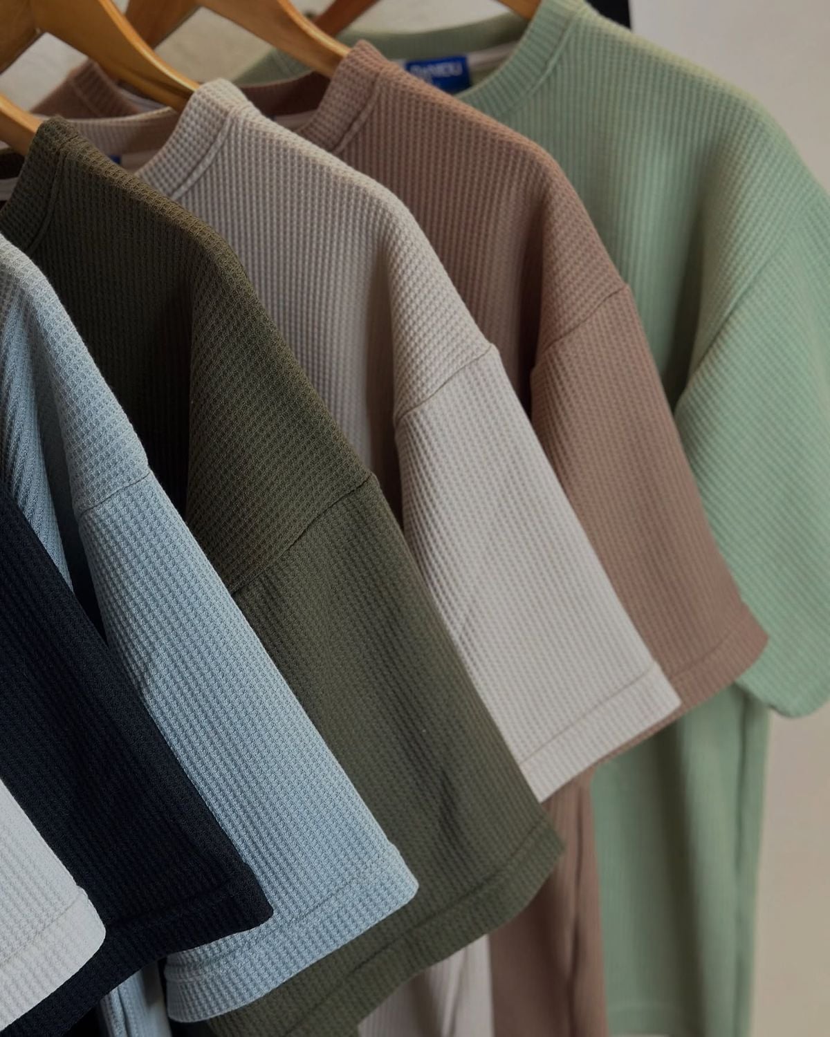 Several ribbed short-sleeve shirts in muted colors—black, light blue, olive green, beige, brown, and sage—hang neatly on wooden hangers, arranged in a row from left to right.