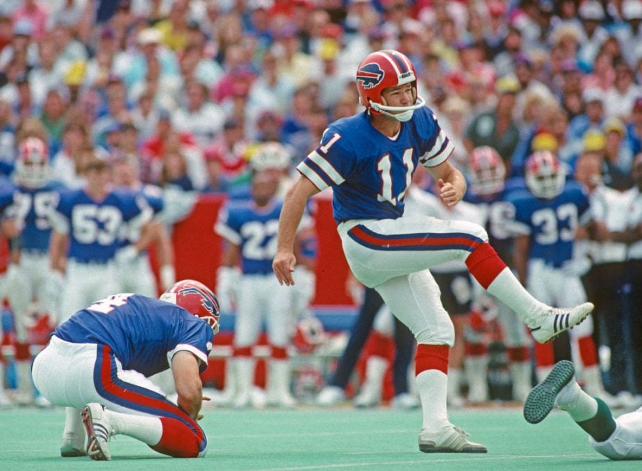 A football player in a blue Buffalo Bills uniform, number 11, kicks a field goal as a teammate holds the ball. Other players and fans watch in the background.