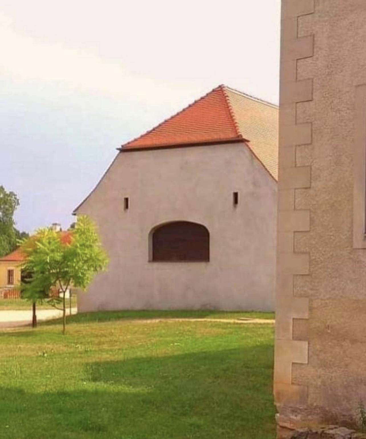 A building with a red-tiled roof and a large, arched window that resembles a smiling face, set on a grassy lawn with a small tree and another building partially visible in the foreground.