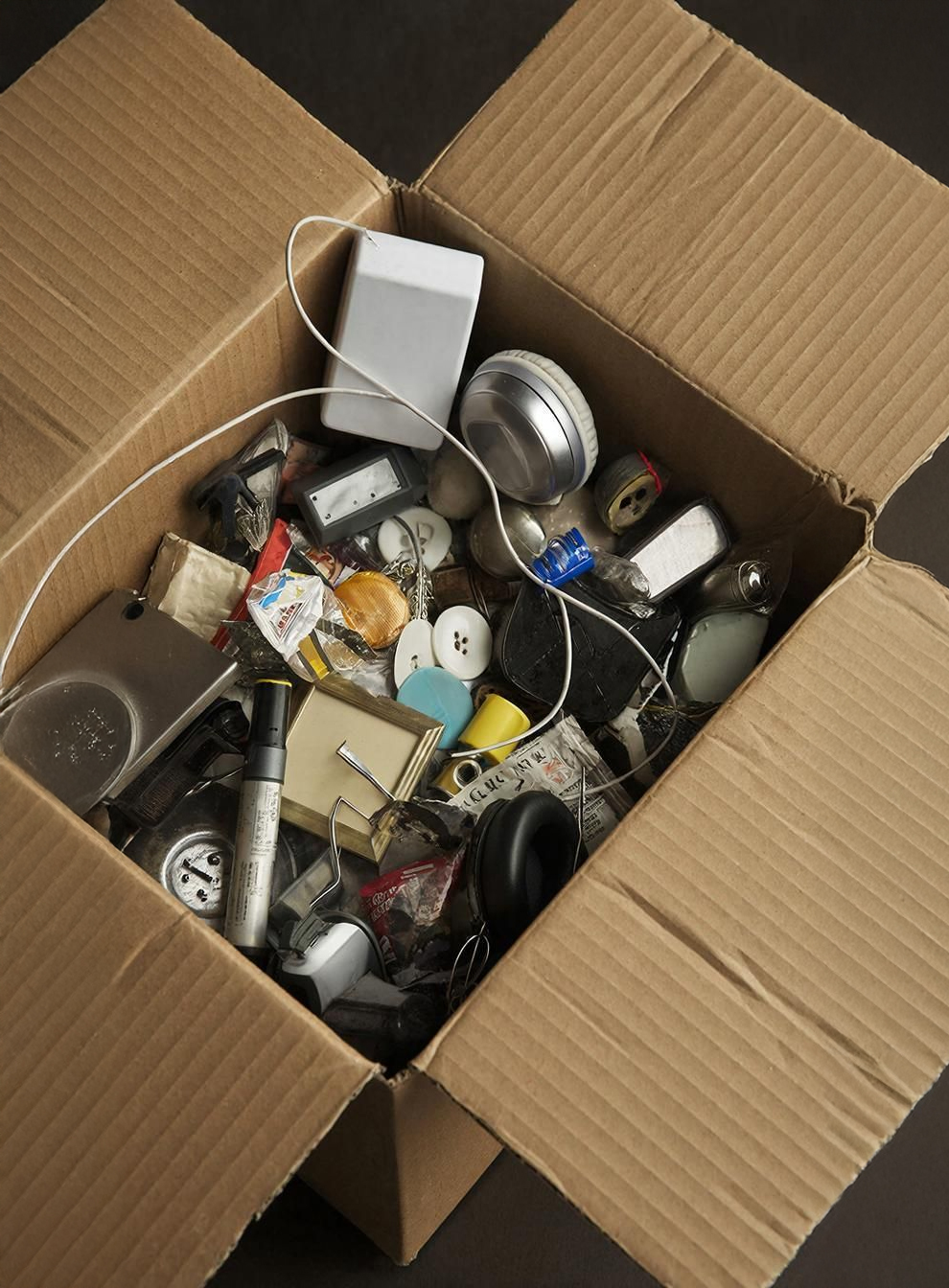An open cardboard box filled with various miscellaneous items, including wires, buttons, a flashlight, a power adapter, batteries, and assorted small gadgets and accessories.