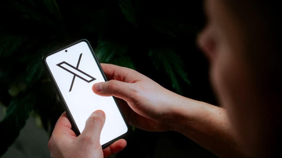 A person holding a smartphone with the X logo, associated with the social media platform formerly known as Twitter, displayed on the screen. The background is out of focus with dark green leaves.