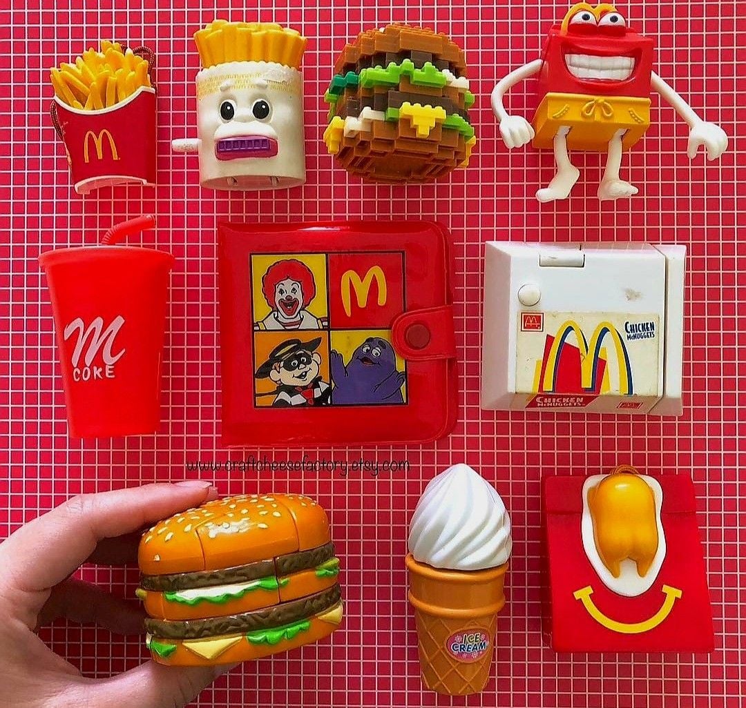 A collection of colorful vintage McDonald's Happy Meal toys is arranged on a red grid background, including burgers, fries, a drink, an ice cream cone, a sauce packet, and character-themed items. A hand holds a toy burger.